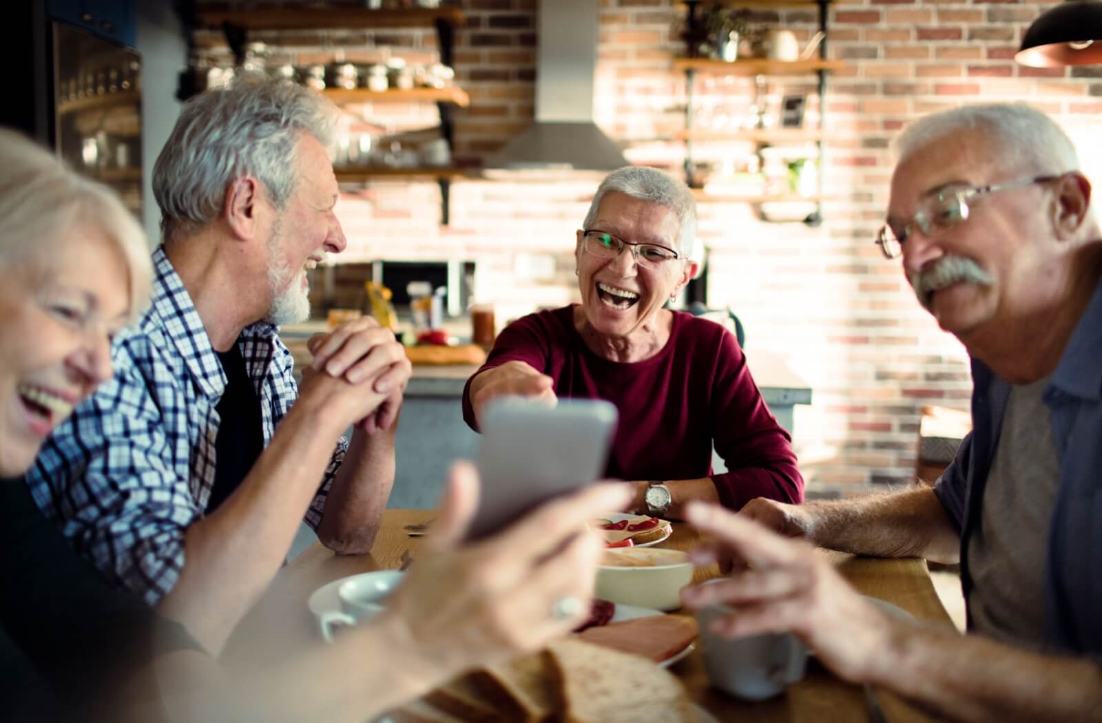 A group of smiling older adults gather around a table, laughing and playing a party game on a smartphone.