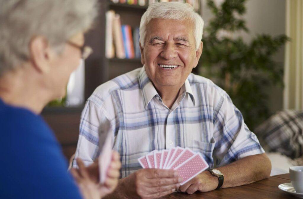 A smiling older adult holds playing cards while sitting at a table with a friend, having fun and a social connection.