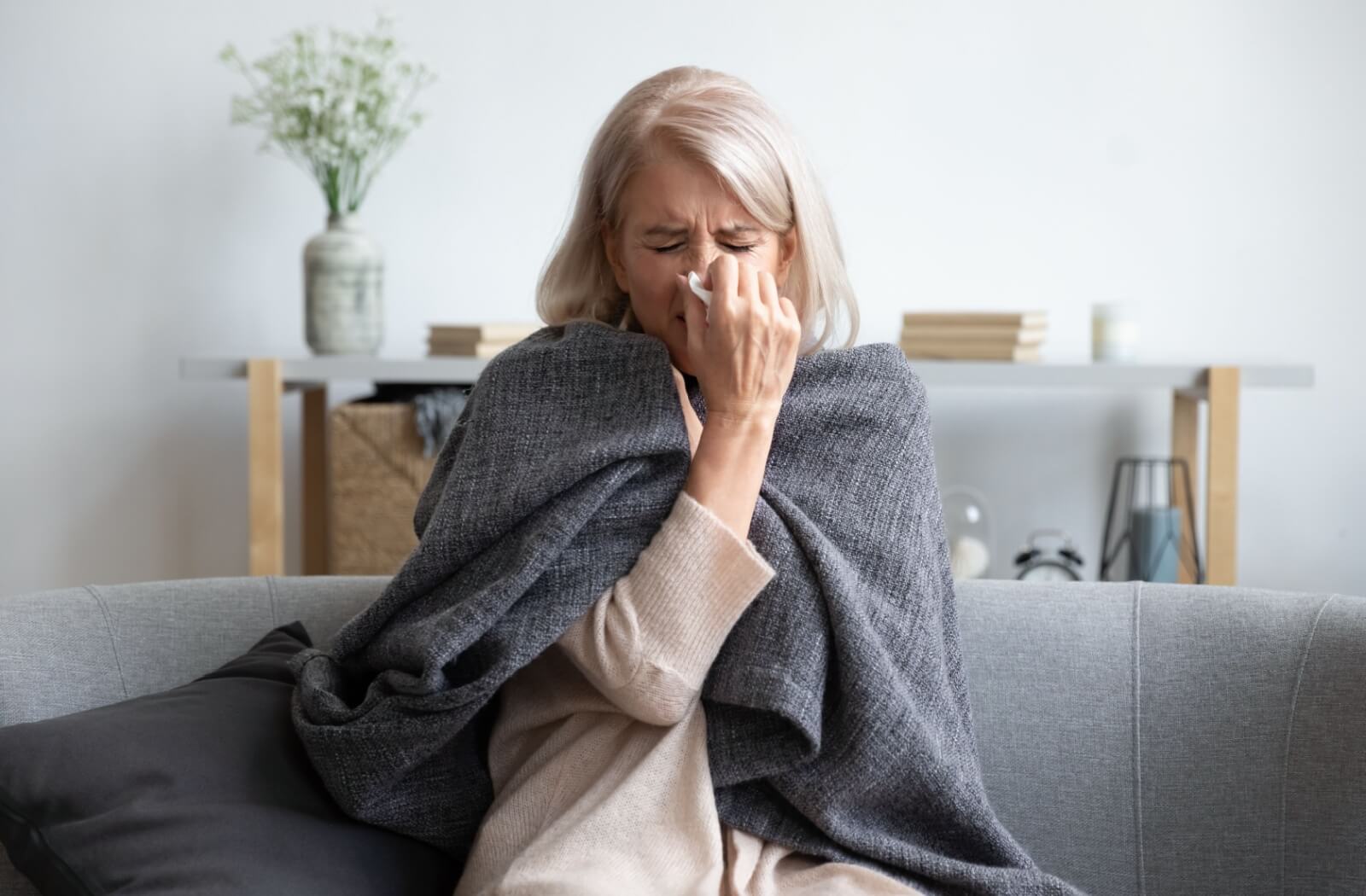 A sick senior sits on a sofa in their living room, covered with a warm blanket, while blowing their nose.