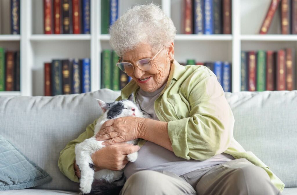 A smiling older adult sits on a couch, gently holding and petting a relaxed black and white cat in independent living.